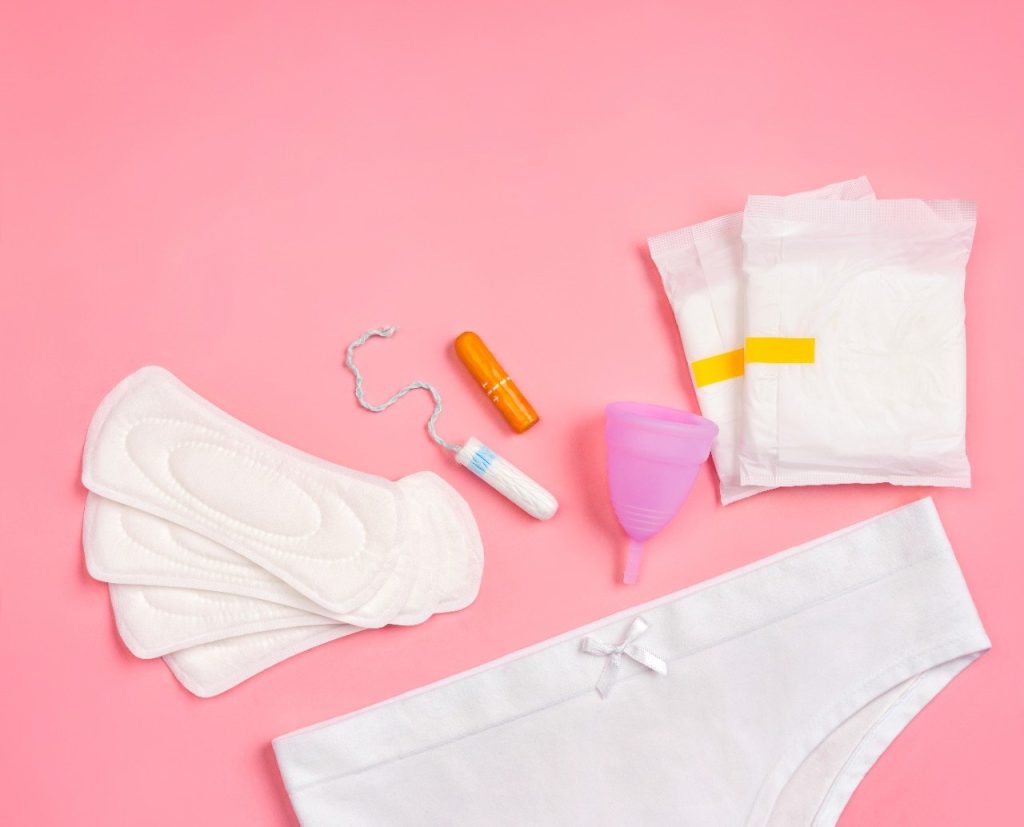 Foto de alguns itens dispostos sob um fundo rosa para falar sobre cuidados com a vagina durante a menstruação.. Um coletor menstrual, alguns absorventes descartáveis e uma calcinha branca.