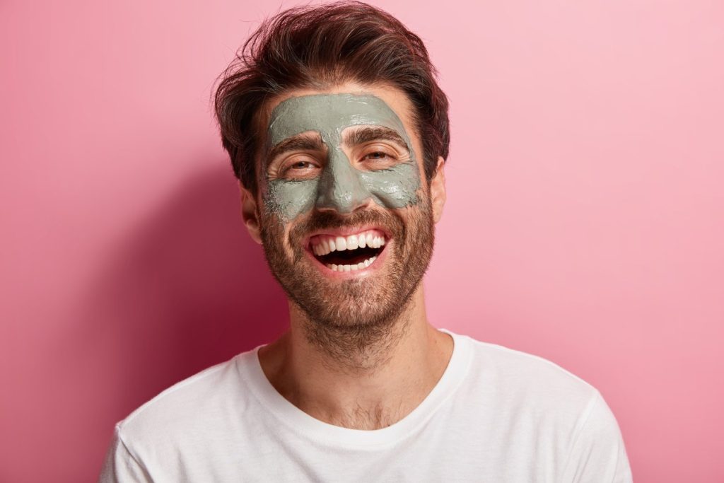 A imagem mostra um homem branco, de cabelos castanhos lisos e barba bem rente ao rosto. Ele está olhando para a câmera e sorri. Em seu rosto, nas áreas da testa, nariz e bochechas, há uma máscara de argila verde. Ao fundo da imagem, é possível ver uma parede rosa. 