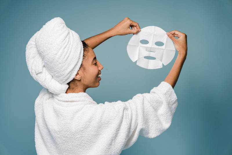 A mulher está de costas para a imagem em um fundo liso e azul, vestindo roupão e toalha na cabeça, ela segura uma máscara facial hidratante com o formato do rosto à sua direita enquanto olha para ela.