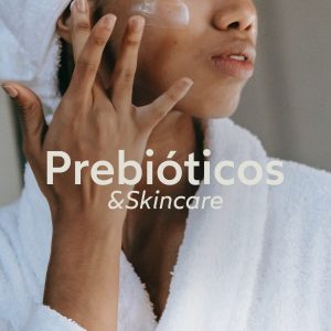 Foto de uma mulher negra pós banho, vestindo um roupão branco e com uma tolha branca nos cabelos passando um creme no rosto. Escrito em branco, no meio da imagem, lê-se “prebióticos & skincare”.