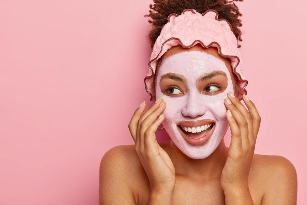 A imagem mostra uma mulher negra de tom claro, de cabelos cacheados avermelhados. O cabelo dela aparece preso com uma faixa rosa. Os ombros dela aparecem nus na foto, pois ela está em um momento de skincare. Em seu rosto, é possível ver uma máscara de argila rosa. Além disso, ela está sorrindo, olhando para o lado esquerdo e suas mãos estão massageando a face. Ao fundo, é possível ver uma parede rosa.