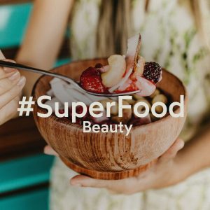 No foco da foto, vemos uma cumbuca de madeira com alguns superalimentos, como frutas vermelhas, castanhas e açaí. Vemos também uma mulher ao fundo, desfocada, segurando a cumbuca e uma colher. Sobre a foto, lê-se em uma cor clara “ #SuperFoodBeauty”.